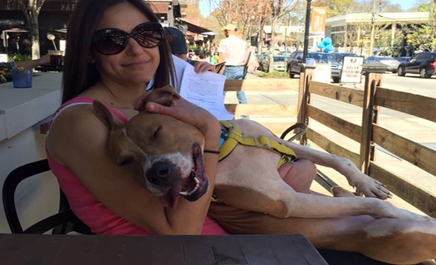 Volunteer, Kimberlie, sitting at a n outdoortable wearing sunglasses with a smiling dog in her lap.