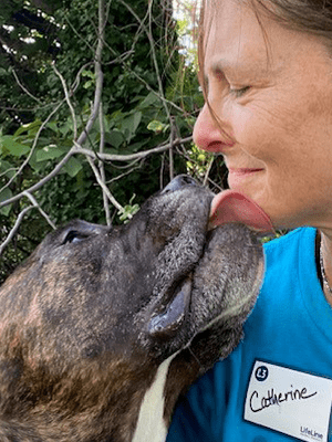 A close up of volunteer, Catherine, knelt down with a brown dog licking her face.