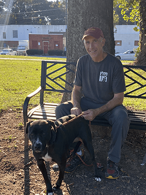 Volunteer, Tom, sitting on a bench outside of the shelter with a black dog standing at his side.